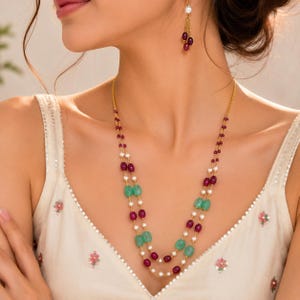 May include: A double-strand gold necklace featuring alternating small white pearls, oval-shaped deep red rubies, and smooth light green emerald beads. A matching gold earring with a single white pearl and a dangling red ruby is also visible.