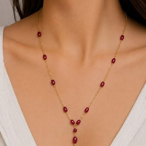 May include: A gold chain necklace featuring evenly spaced, small, oval-shaped deep red beads. The necklace has a Y-shaped drop design at the center, with a single vertical strand ending in a bead, creating a delicate and symmetrical appearance.