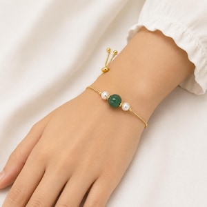 May include: A gold-toned adjustable chain bracelet featuring a central ribbed dark green bead flanked by two small white spherical beads. The bracelet includes a sliding mechanism with two small gold-toned beads at the end of the chain.