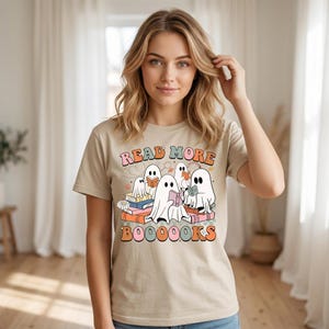 May include: A beige short-sleeve t-shirt featuring a graphic of five white ghosts reading books. The text reads READ MORE BOOOOOKS in a retro font. The design includes colorful books, small stars, and flowers in shades of orange, pink, and blue.