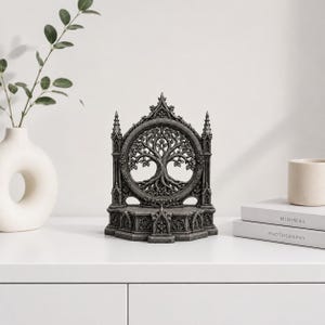 Puede incluir: Una escultura decorativa de color gris oscuro, similar a la piedra, que presenta un intrincado diseño de árbol de la vida dentro de un marco circular. La estructura se apoya en una base rectangular con detalles arquitectónicos góticos y agujas.
