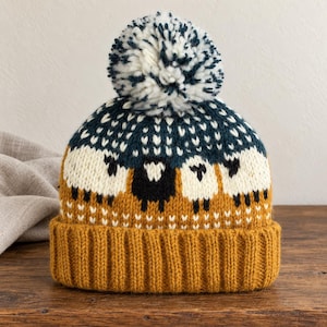 Könnte beinhalten: Eine Strickmütze mit einem wiederkehrenden Muster aus weißen und schwarzen Schafen auf dunkelblau-grünem Hintergrund. Die Mütze hat einen breiten, gerippten Umschlag in Senfgelb und ist mit einem großen, flauschigen Bommel aus weißem und blau-grünem Garn versehen.