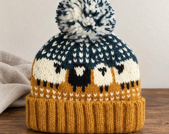 Puede incluir: Un gorro de punto con un patrón repetitivo de ovejas blancas y negras sobre un fondo verde azulado oscuro. El gorro tiene un borde ancho acanalado de color amarillo mostaza en la base y está rematado con un pompón grande y esponjoso de lana blanca y azulada.