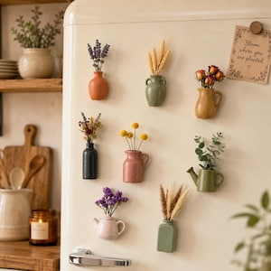 Könnte beinhalten: Eine Sammlung von acht kleinen magnetischen Keramikvasen an einem Kühlschrank, die jeweils mit Trockenblumen oder Grünpflanzen bestückt sind. Die Vasen variieren in ihrer Form, darunter Krüge, Kannen und Töpfe in Farben wie Terrakotta, Salbeigrün und Schwarz.
