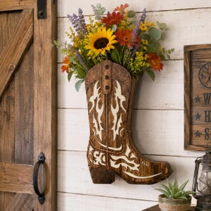 Può includere: Un vaso da parete a forma di stivale da cowboy realizzato in legno marrone scuro con intagli decorativi bianchi. Lo stivale è riempito con una vivace composizione floreale artificiale con un grande girasole giallo, margherite rosse e lavanda.