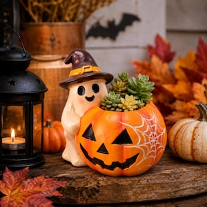 May include: A ceramic planter shaped like a smiling ghost wearing a brown witch hat, hugging an orange jack-o'-lantern pumpkin with a white spiderweb design. The pumpkin contains a small green succulent plant with various shades of foliage.