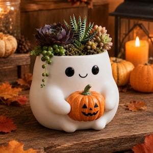 Pode incluir: Um vaso de cerâmica branca em forma de fantasma que segura uma pequena abóbora laranja. O fantasma tem dois olhos pretos circulares e uma boca curva simples. O vaso contém uma variedade de suculentas verdes e roxas escuras.