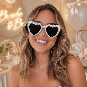 May include: Heart-shaped sunglasses with white pearl-like embellishments along the frames. The left lens features the text BRIDE and the right lens features the text TO BE in black capital letters. The lenses are dark and opaque in colour.