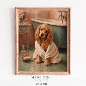 May include: A framed wall art print depicting a golden-brown spaniel dog wrapped in a beige towel, sitting on a green tiled floor in a bathroom. A vintage bathtub, a small wooden side table, a glass perfume bottle, and a soap dish are visible.