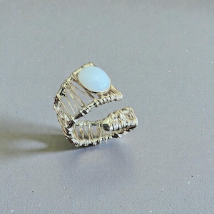 Peut inclure: Bague filaire argentée avec une pierre ovale bleu pâle. La bague a un design ouvert, avec la pierre précieuse sertie en haut. Le fil métallique crée un aspect texturé en forme de cage.