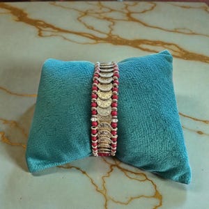 Peut inclure: Un bracelet de pièces de monnaie dorées avec des perles rouges, présenté sur un coussin en velours turquoise. Le bracelet est composé de plusieurs breloques dorées en forme de pièces et de perles rouges.