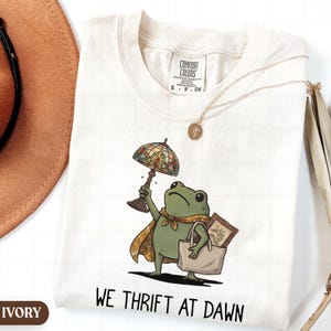 May include: An ivory t-shirt featuring a graphic of a green frog wearing a patterned scarf and cape, holding a stained glass lamp and a tote bag containing a framed picture. The text "WE THRIFT AT DAWN" is printed below the frog illustration.