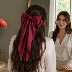 Peut inclure: Un nœud pour cheveux bordeaux profond fabriqué dans un tissu satiné lisse. L'accessoire présente un nœud structuré sur le dessus avec deux longs pans fluides qui tombent le long des cheveux, créant un aspect classique et élégant.