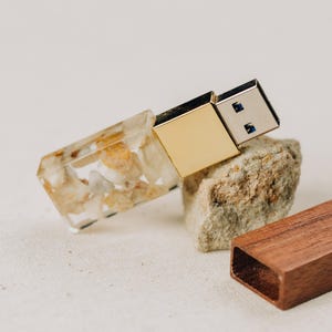 Może przedstawiać: Pamięć USB z korpusem z przezroczystej żywicy z zatopionymi małymi białymi i beżowymi kamykami, złotym metalowym złączem oraz oddzielną prostokątną ciemnobrązową drewnianą nasadką leżącą obok na teksturowanej powierzchni.