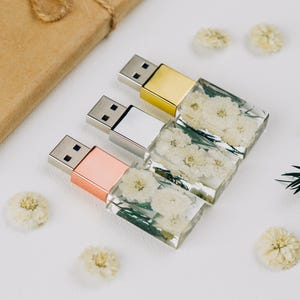 Puede incluir: Tres memorias USB con cuerpos rectangulares de resina transparente que contienen pequeñas flores blancas secas. Cada unidad tiene una tapa metálica con acabado en oro rosa, plata u oro, y conectores USB-A estándar en los extremos.