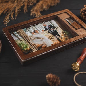 Op de afbeelding: Een rechthoekige donkerbruine houten doos met een scharnierend deksel voorzien van een glazen venster waarin een foto van een bruidspaar te zien is. De doos bevat een speciaal vakje voor een bijpassende houten USB-stick.