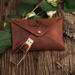 May include: A brown leather envelope-style pouch with a gold-tone snap closure and the text Wedding Day engraved above a sprig illustration, accompanied by a clear and gold-tone USB flash drive with a rectangular transparent body.
