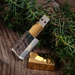 Può includere: Una chiavetta USB con un corpo in cristallo trasparente rettangolare e un connettore in metallo color oro lucido. Il dispositivo include un tappo rettangolare in metallo color oro abbinato che poggia accanto all'unità principale su una superficie in legno.