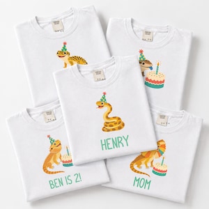 Könnte beinhalten: Fünf weiße T-Shirts mit bunten Illustrationen von Reptilien mit Partyhüten. Die Motive zeigen einen Leopardgecko, eine Schlange und Bartagamen, teilweise mit Geburtstagskuchen. Der Text umfasst "BEN IS 2!", "HENRY" und "MOM".