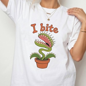 Puede incluir: Una camiseta blanca de manga corta con un gráfico de una planta carnívora Venus atrapamoscas verde en una maceta de color terracota. Sobre la planta, el texto "I bite" está impreso en una fuente naranja, redondeada y negrita con un borde oscuro.