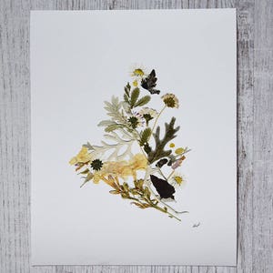 May include: A pressed flower art print featuring a variety of dried flowers and leaves arranged in a triangular shape on a white background. The flowers are mostly white and yellow, with some green leaves and a few brown accents.