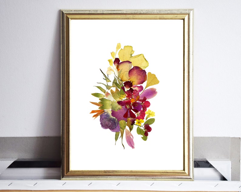 A framed print of a pressed flower arrangement. The flowers are arranged in a bouquet shape and include yellow, red, purple, and green tones. The flowers are pressed against a white background.