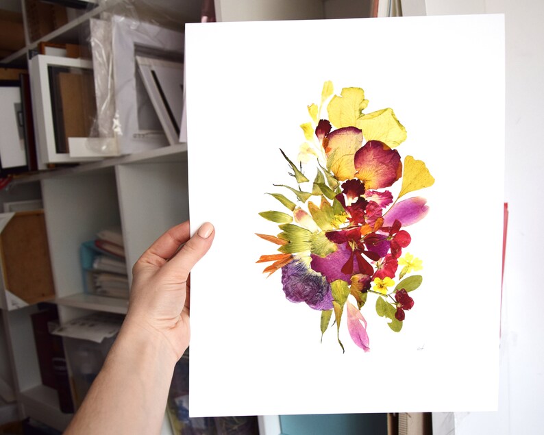 A pressed flower art print with a white background. The print features a bouquet of yellow, purple, red, and pink flowers. The flowers are arranged in a loose, organic shape. The print is signed in the bottom right corner.