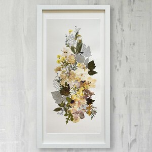 May include: A framed pressed flower art piece with a white frame and a white mat. The flowers are arranged in a vertical bouquet and are mostly yellow and cream with some brown and green accents.