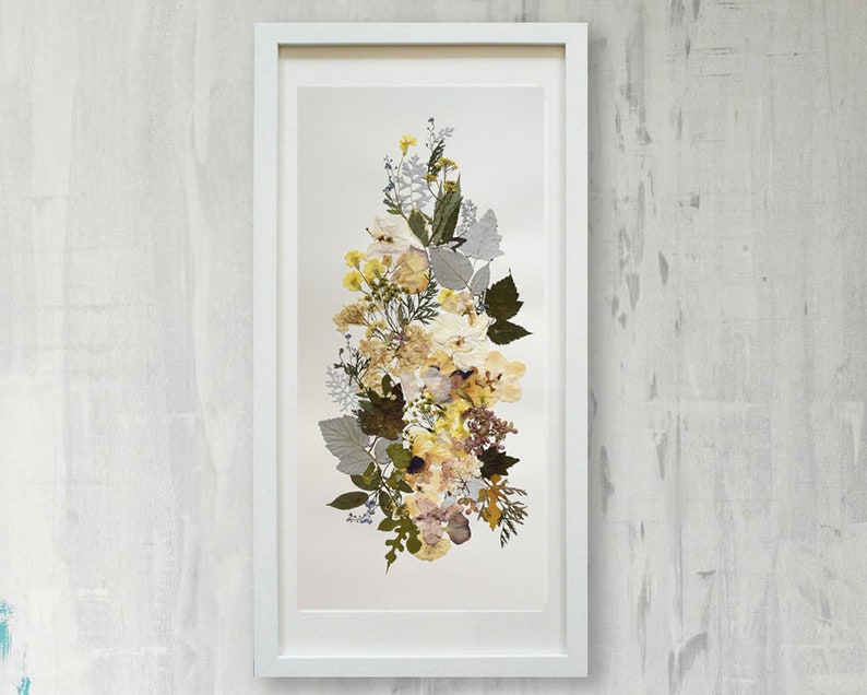 May include: A framed pressed flower art piece with a white frame and a white mat. The flowers are arranged in a vertical bouquet and are mostly yellow and cream with some brown and green accents.
