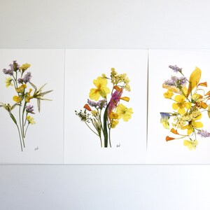 Puede incluir: Tres impresiones enmarcadas de flores prensadas, cada una con una disposici&oacute;n diferente de flores amarillas, moradas y naranjas.