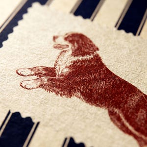 Puede incluir: Una tarjeta rectangular con borde dentado que presenta una ilustraci&oacute;n de color rojo oscuro de un perro en posici&oacute;n de salto. La tarjeta tiene una superficie texturizada de color blanco roto sobre un fondo de rayas verticales azul oscuro y crema.