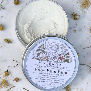 May include: Open tin of baby diaper rash cream with the text "Baby Bum Bum" and "Maternal Botanica". The cream is white and surrounded by dried flowers and herbs. The tin is 2 oz / 57g.