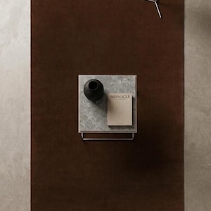 May include: A large rectangular dark brown area rug featuring a centered square side table with a grey marble top. On the table sits a black vase and a tan book with the word MONOCLE printed on the cover. A white spherical floor lamp stands nearby.