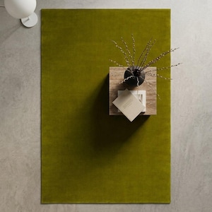 May include: A rectangular olive green area rug with a smooth, solid-colored surface. The rug is positioned on a light gray floor, featuring a square stone side table topped with a dark vase containing thin branches and a small, light-colored book.