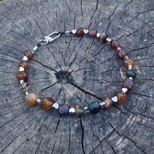 May include: A bracelet featuring polished, multi-colored tumbled stones in shades of brown, orange, and dark green, interspersed with small silver-toned metal spacers. The stones are strung on a dark cord and secured with a silver-toned lobster clasp.