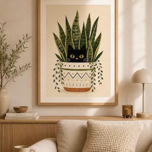 Op de afbeelding: Een ingelijste kunstdruk met een zwarte kat die uit een pot met slangengras kijkt. De kat heeft gele ogen. De pot is wit met een geometrisch ontwerp en groene ranken. Het kunstwerk wordt tentoongesteld in een kamer met neutrale tinten.