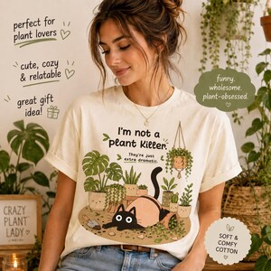 May include: A cream-colored t-shirt featuring a graphic of a black cat lying on the ground surrounded by various potted plants. The text reads: I'm not a plant Killer. They're just extra dramatic. The design includes a small trowel and a hanging plant.