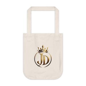 May include: A natural-colored canvas tote bag featuring a centered graphic design. The design consists of the letters JD in a bold, metallic gold font, enclosed within a circular gold frame and topped with a detailed gold crown illustration.
