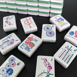 May include: A set of rectangular mahjong tiles featuring a three-layered design with a green top, white middle, and pink bottom. The white faces display colorful engraved illustrations including birds, umbrellas, pagodas, and floral motifs with numbers.
