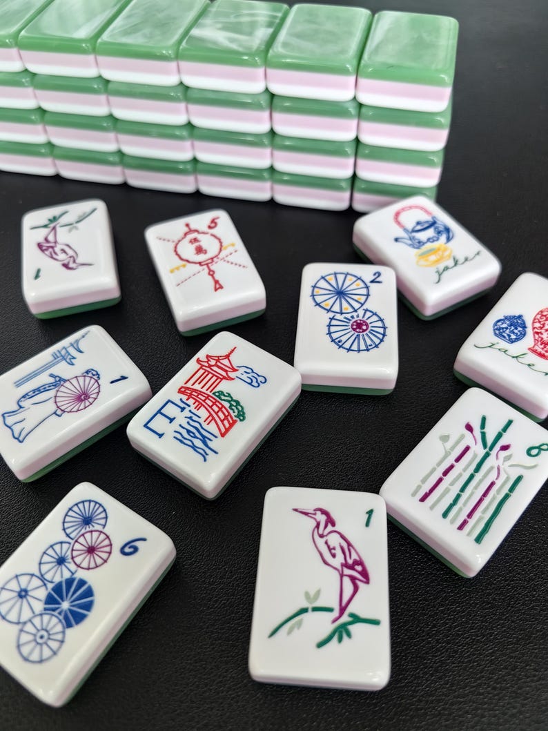 May include: A set of rectangular mahjong tiles featuring a three-layered design with a green top, white middle, and pink bottom. The white faces display colorful engraved illustrations including birds, umbrellas, pagodas, and floral motifs with numbers.