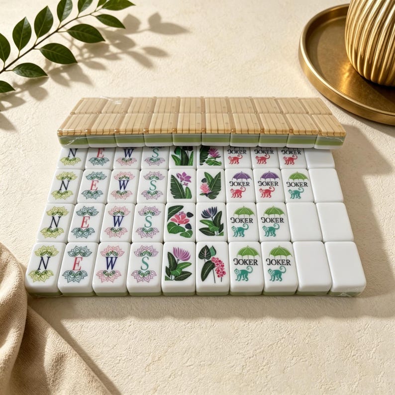 Puede incluir: Un conjunto de fichas de mahjong rectangulares dispuestas en filas. Las fichas presentan ilustraciones coloridas de flores, hojas y monos bajo paraguas con el texto JOKER, junto a fichas que muestran las letras N, E, W y S en marcos florales.
