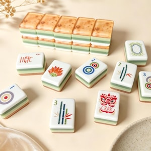 May include: A set of rectangular mahjong tiles featuring a white top surface with colorful illustrations including a lotus, bamboo, circles, and a lion mask with the text CRAK. The tiles have a layered design with green and tan sides.