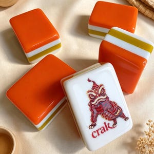 K&ouml;nnte beinhalten: Vier rechteckige Mahjong-Spielsteine mit orangefarbenen, wei&szlig;en und goldenen Schichten an den Seiten. Ein Spielstein zeigt eine detaillierte rot-braune Illustration eines L&ouml;went&auml;nzers und den roten Schriftzug "crak" auf der wei&szlig;en Vorderseite.
