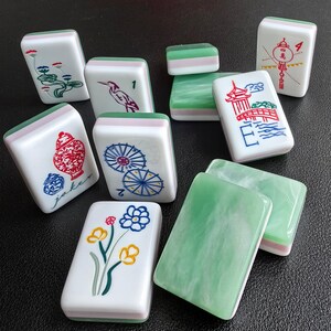 May include: A collection of rectangular Mahjong tiles featuring white faces with colorful illustrations including flowers, a bird, a bridge, a vase, and umbrellas, alongside solid green marbled backs and layered white and green side panels.