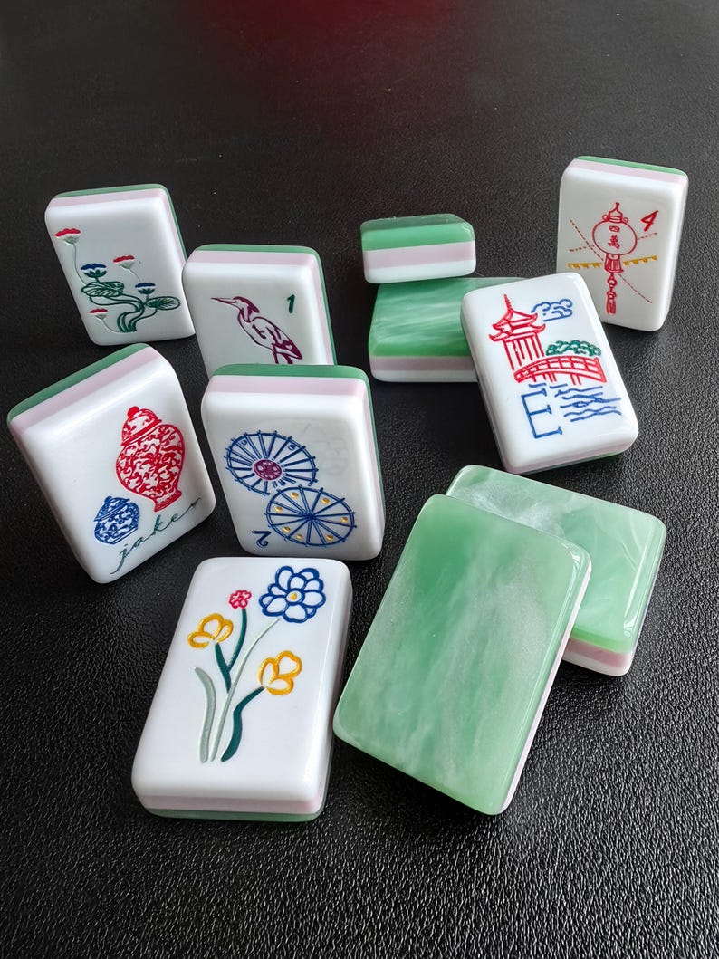 May include: A collection of rectangular Mahjong tiles featuring white faces with colorful illustrations including flowers, a bird, a bridge, a vase, and umbrellas, alongside solid green marbled backs and layered white and green side panels.