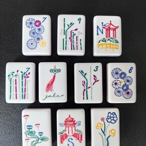May include: A set of ten white rectangular mahjong tiles featuring colorful engraved designs. The tiles display various illustrations including bamboo stalks, pagodas, flowers, umbrellas, and a red tassel with the text jaker and the letter S.