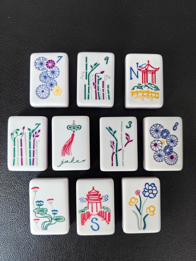 May include: A set of ten white rectangular mahjong tiles featuring colorful engraved designs. The tiles display various illustrations including bamboo stalks, pagodas, flowers, umbrellas, and a red tassel with the text jaker and the letter S.