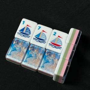 K&ouml;nnte beinhalten: Drei rechteckige Mahjong-Spielsteine mit einem wei&szlig;en oberen Bereich, der eine blaue Segelbootgrafik und die Zahlen 1, 7 und 2 zeigt, kombiniert mit einem unteren Bereich in blau-goldener Marmoroptik, neben einem vertikalen Stein mit gr&uuml;n-rosa Streifen.