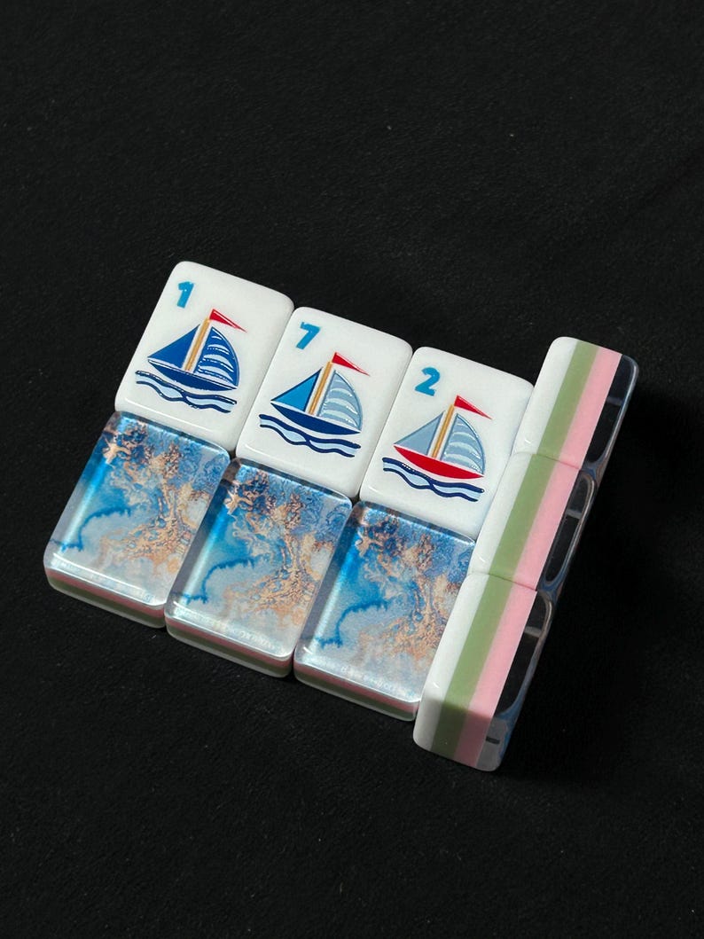 K&ouml;nnte beinhalten: Drei rechteckige Mahjong-Spielsteine mit einem wei&szlig;en oberen Bereich, der eine blaue Segelbootgrafik und die Zahlen 1, 7 und 2 zeigt, kombiniert mit einem unteren Bereich in blau-goldener Marmoroptik, neben einem vertikalen Stein mit gr&uuml;n-rosa Streifen.