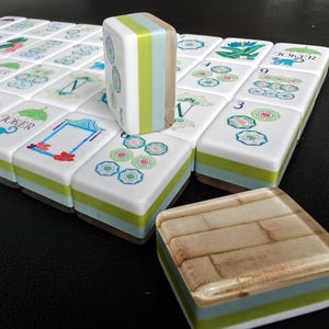 Puede incluir: Un juego de fichas de Mahjong rectangulares con caras blancas que presentan ilustraciones coloridas de flores, paraguas y la palabra JOKER. Las fichas tienen lados en capas de color verde, azul claro y madera, con una ficha vertical y otra plana.